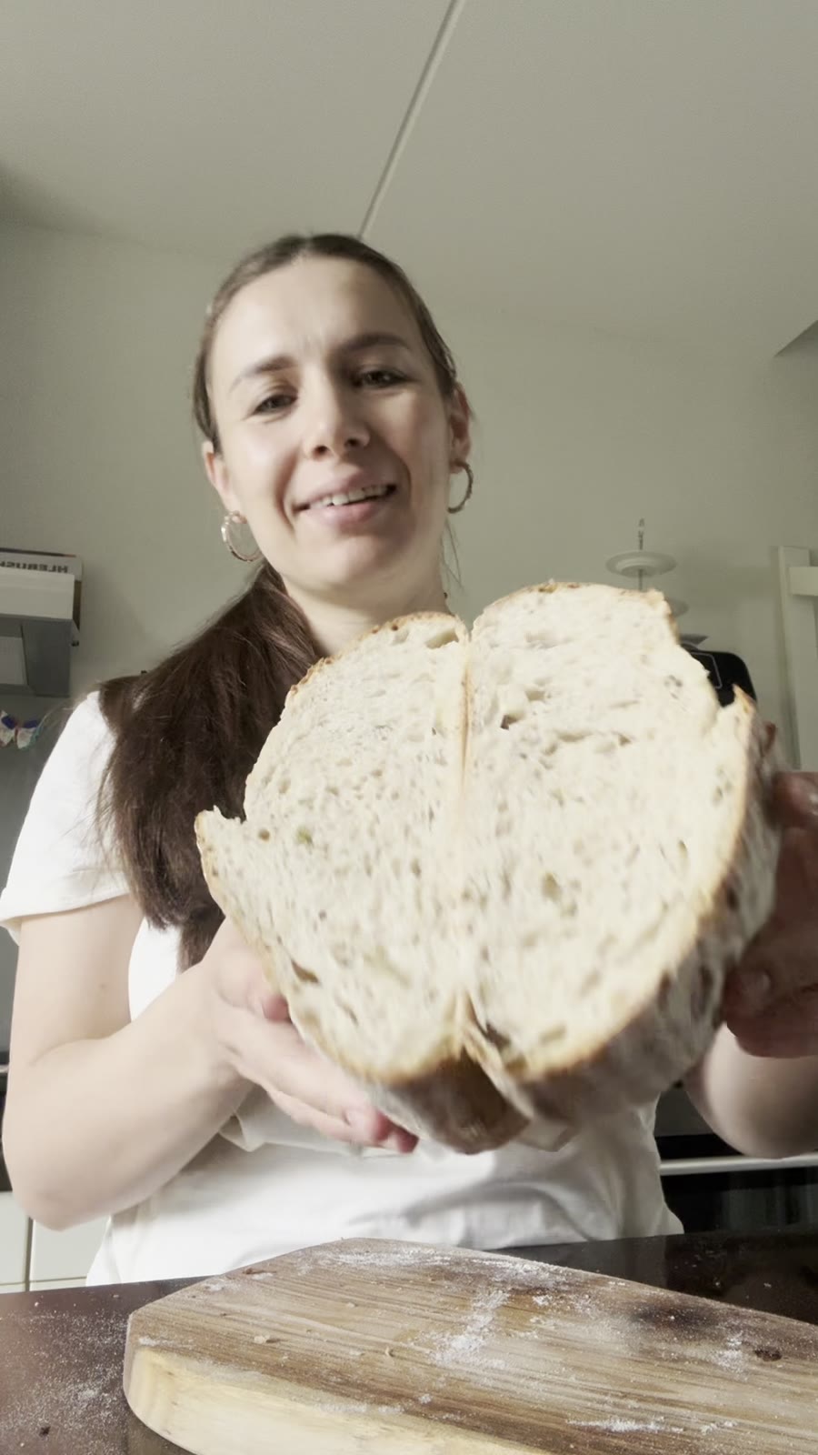 Ana holding two halves of a freshly baked scored country sourdough loaf over a wooden board in her kitchen